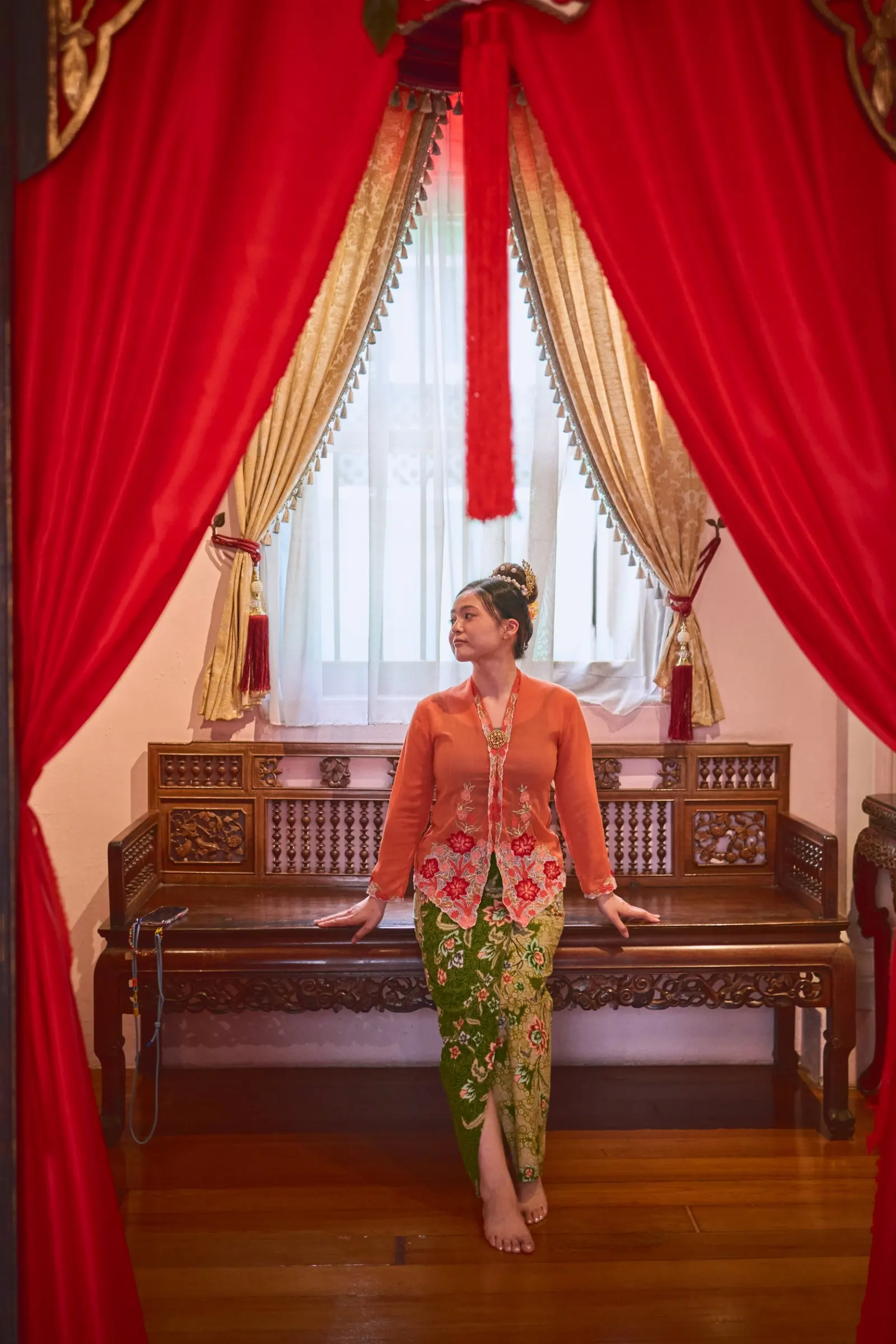 Woman in an orange Nyonya kebaya and green batik sarong sitting on a carved wooden bench between red curtains, Penang