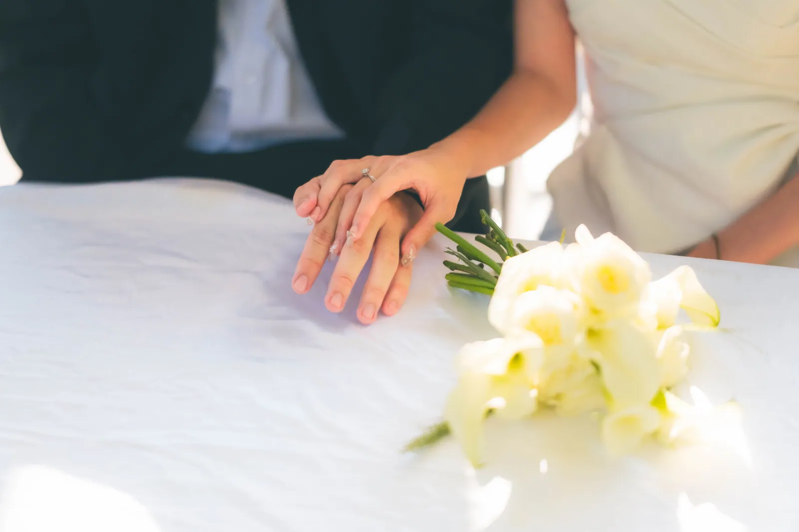 Bride's hand with an engagement ring resting on the groom's hand beside a white bouquet