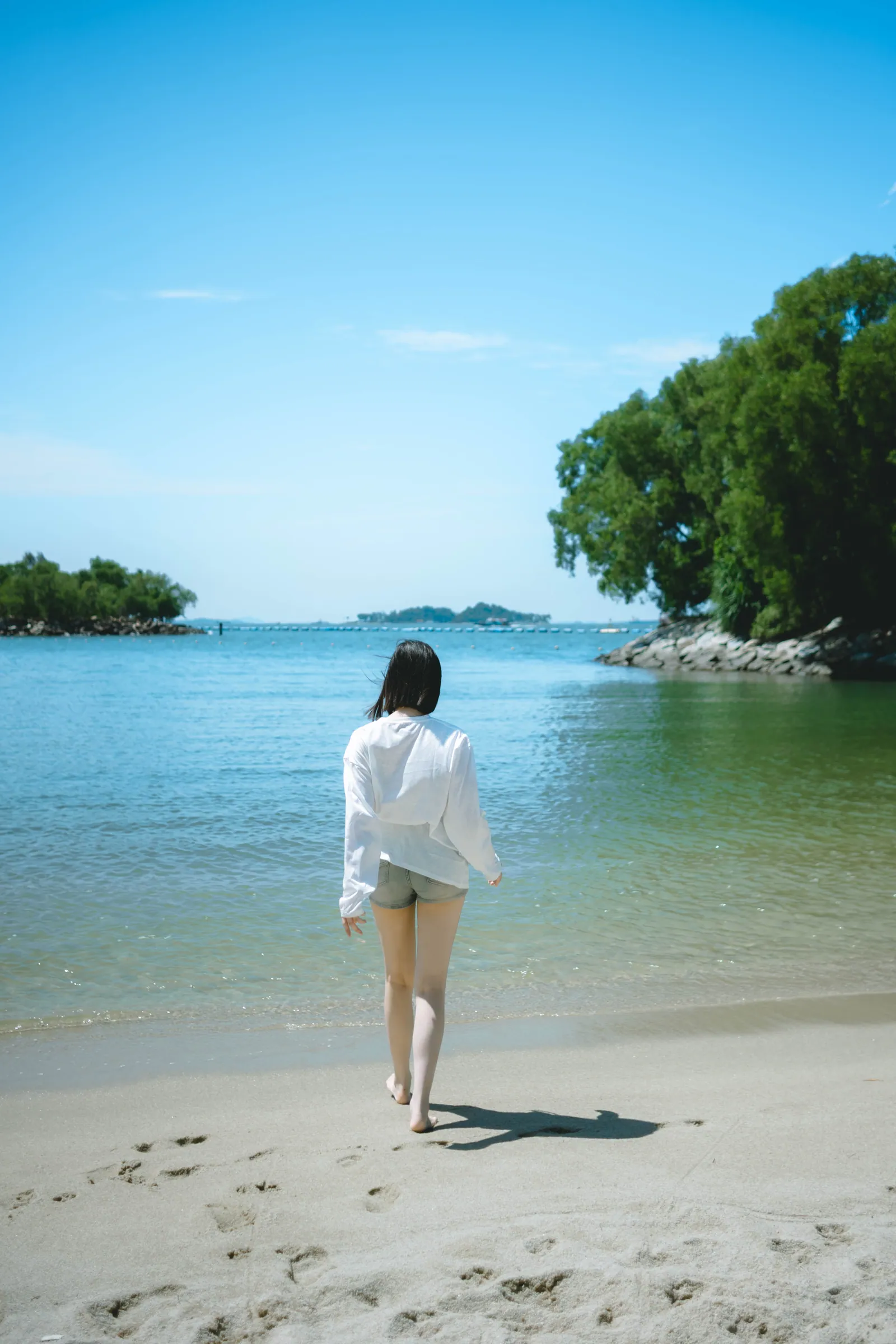 穿宽松白衬衫的女生走进平静的蓝色海水,远处是一座绿树小岛,新加坡圣淘沙海滩