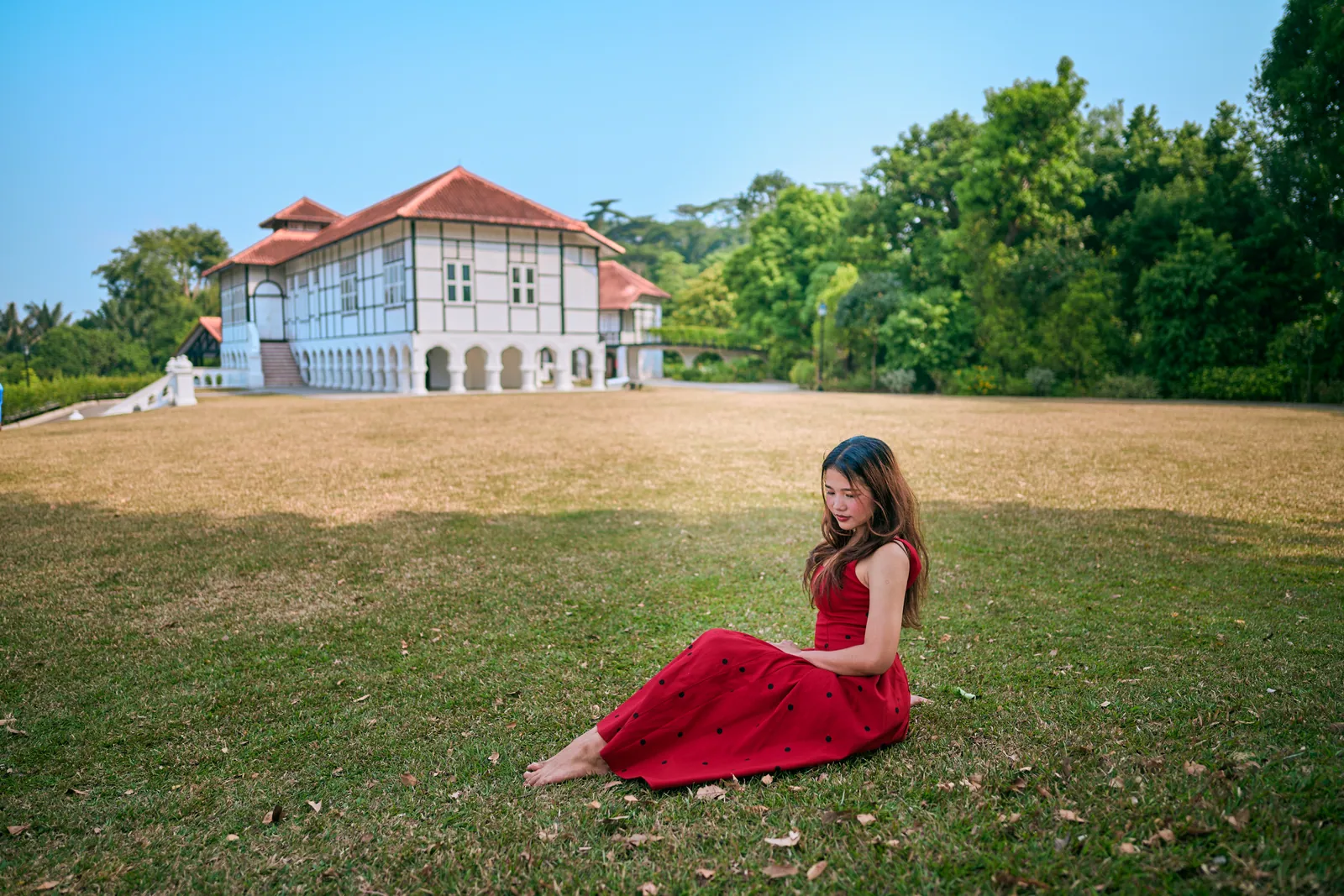 Woman in a red polka-dot dress sitting barefoot on a lawn, with the black-and-white colonial Burkill Hall behind, Singapore Botanic Gardens
