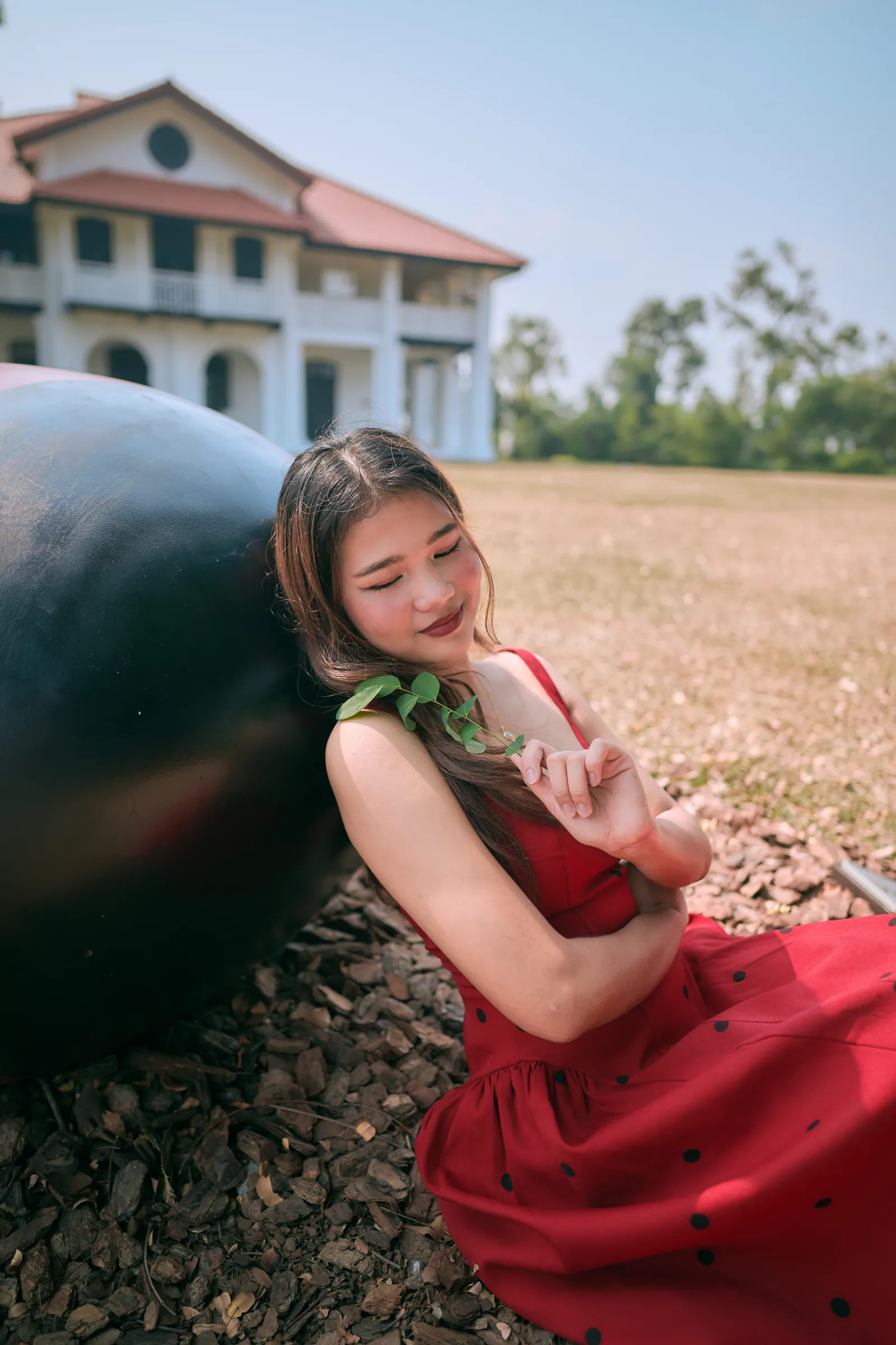 Woman in a red polka-dot dress leaning against a dark sculpture with her eyes closed, holding a green sprig, with a white colonial house behind, Singapore Botanic Gardens