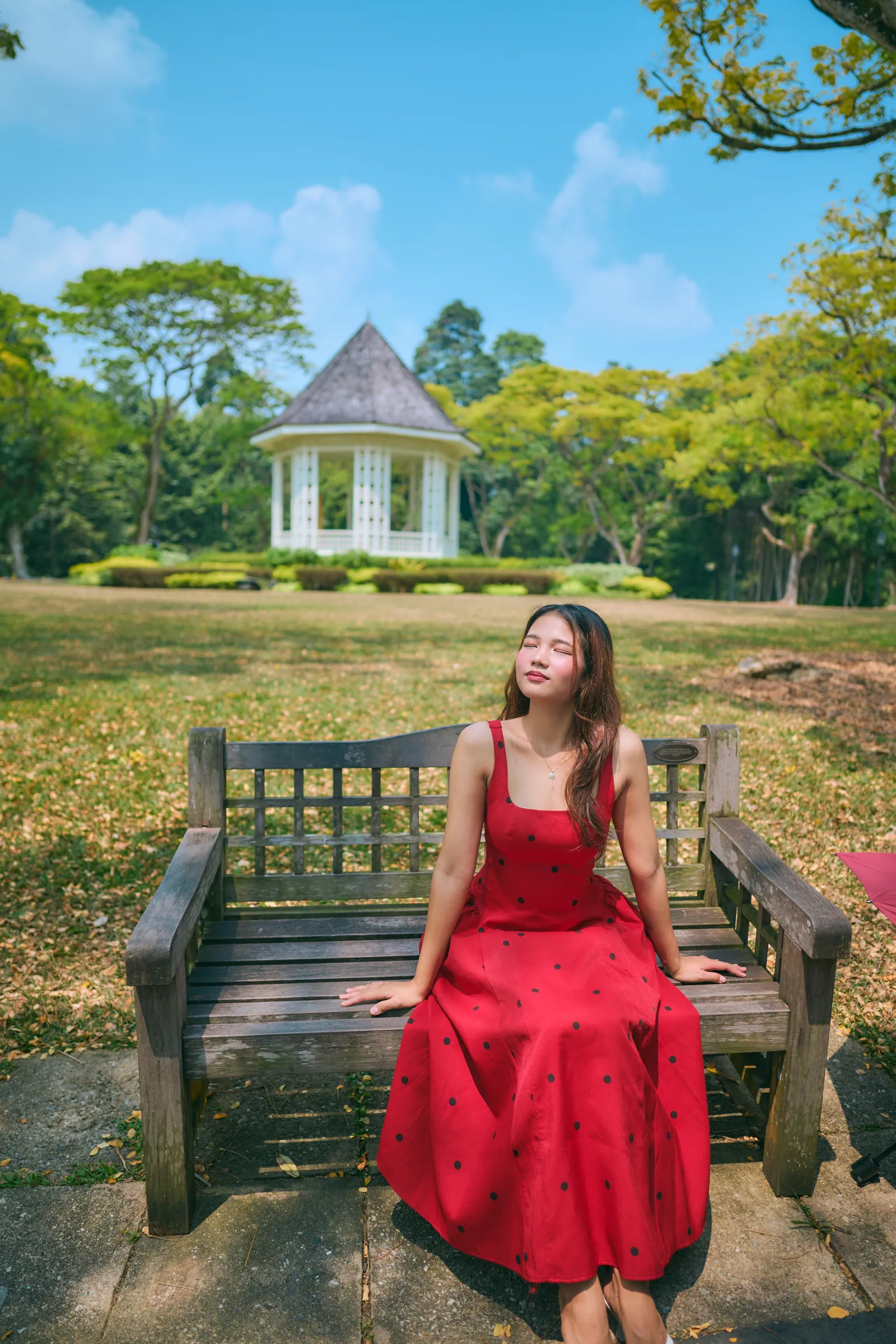 新加坡植物园旅拍:穿红色波点连衣裙的女生闭眼坐在木长椅上晒太阳,身后是白色音乐亭
