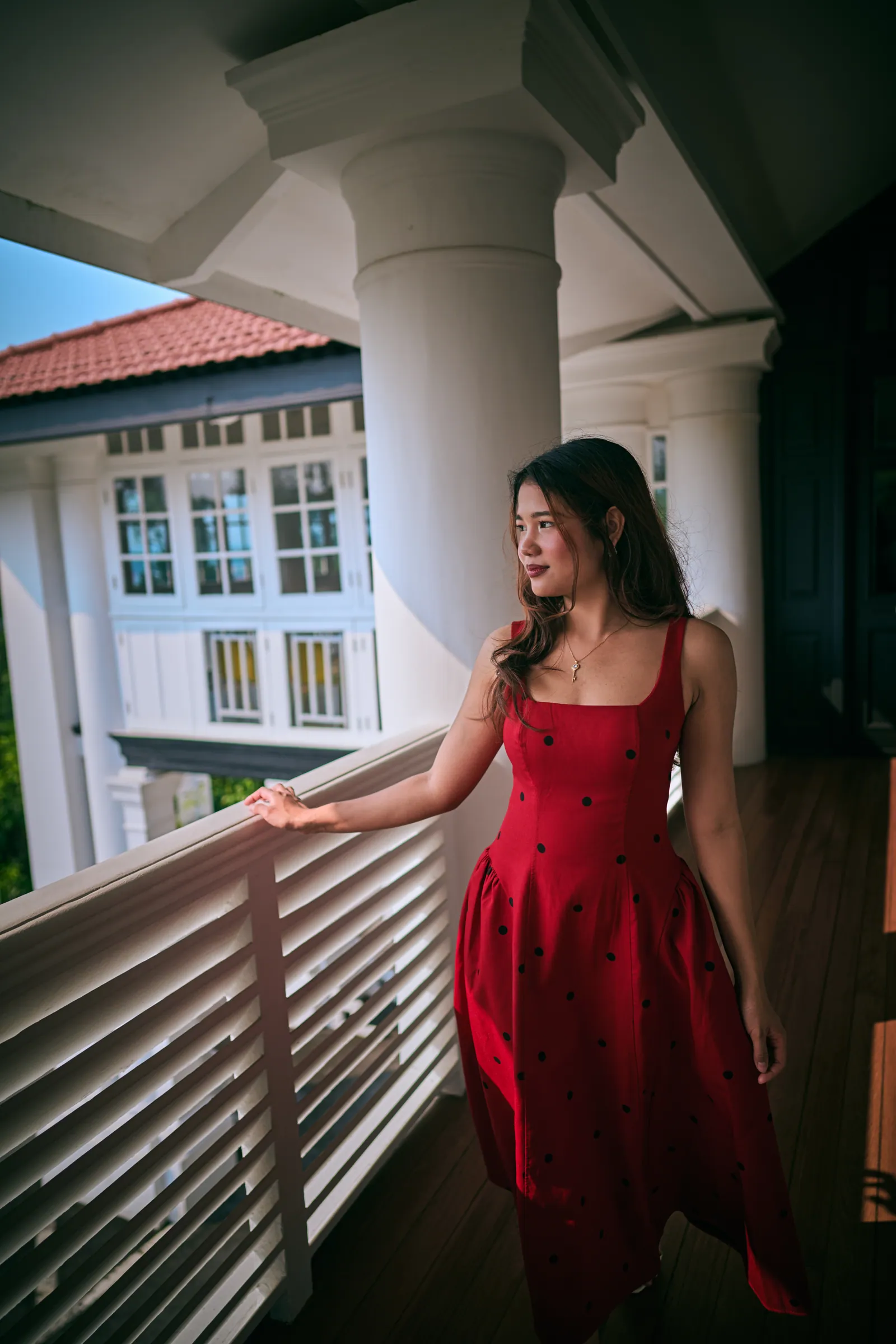Woman in a red polka-dot dress standing on a colonial veranda with white columns and louvred railings, looking out, Singapore Botanic Gardens