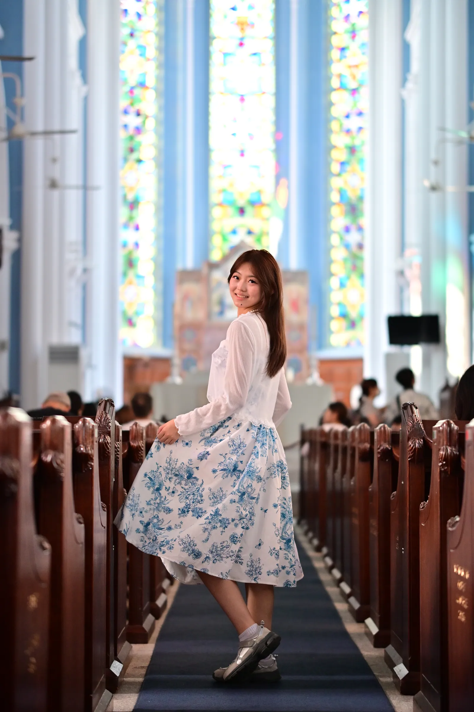 Woman in a white cardigan and blue floral skirt turning to smile in the aisle of St Andrew's Cathedral, with stained glass windows behind, Singapore church photoshoot