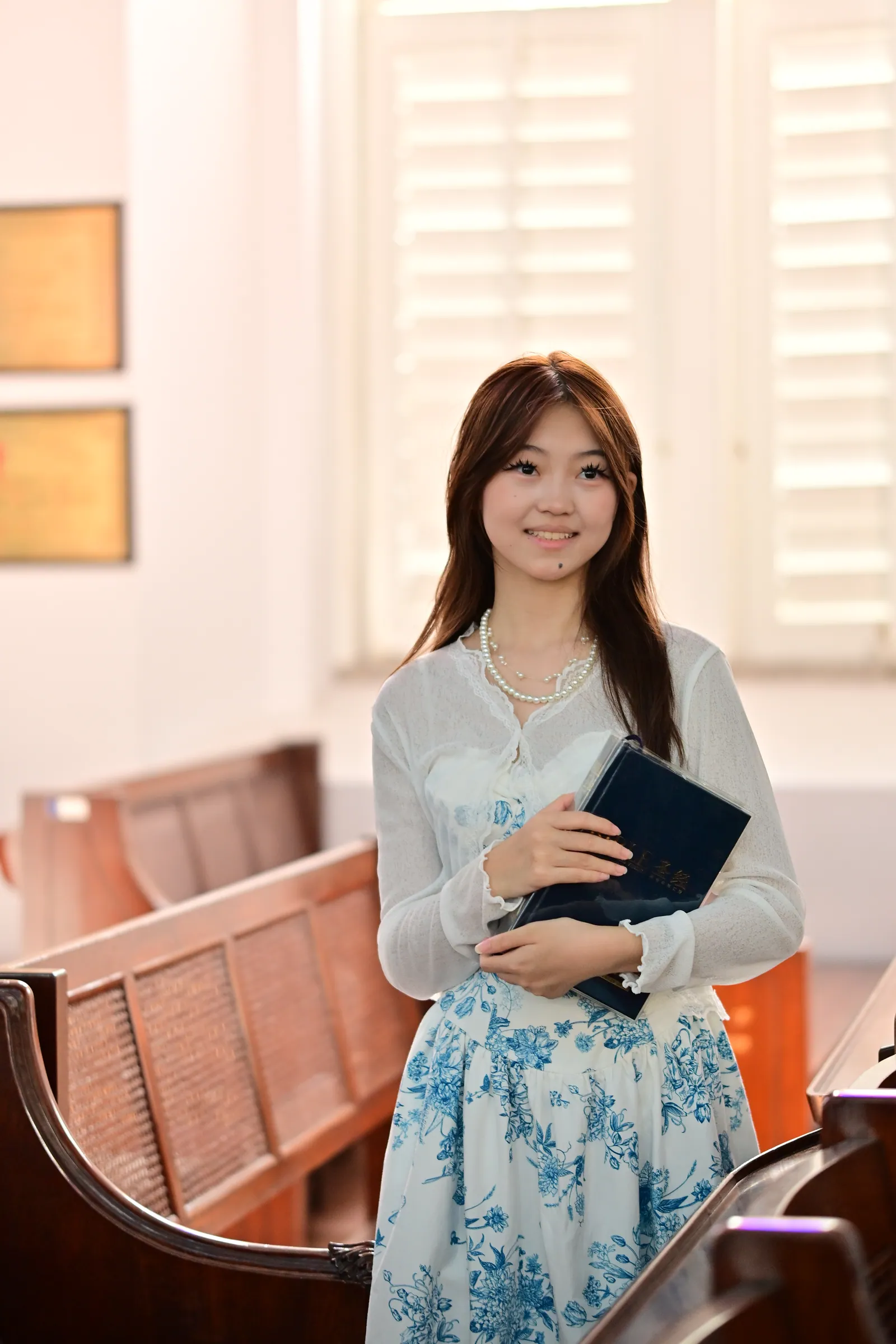 Woman in a white cardigan and blue floral skirt holding a book and smiling, standing between rows of wooden pews by bright louvred windows, St Andrew's Cathedral