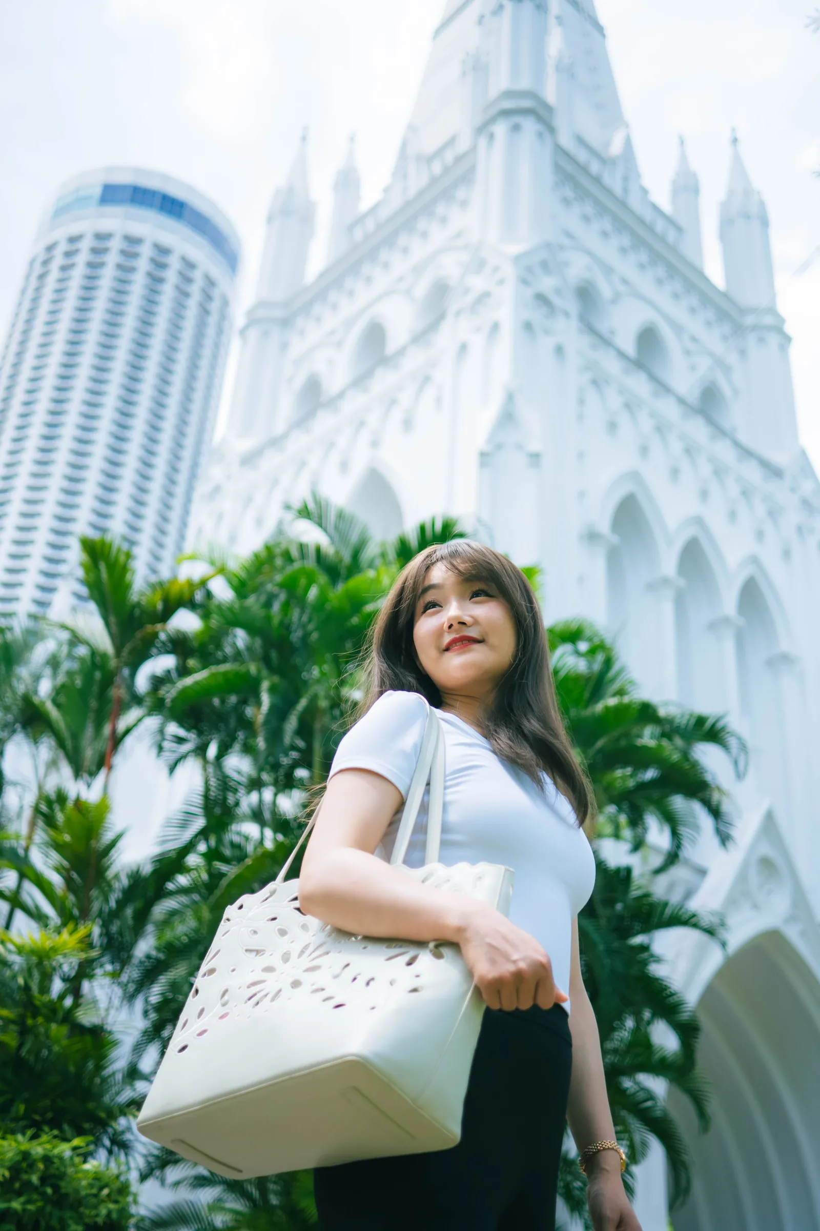 Woman with a white tote bag smiling in front of palms and the white Gothic tower of St Andrew's Cathedral, shot from a low angle, Singapore