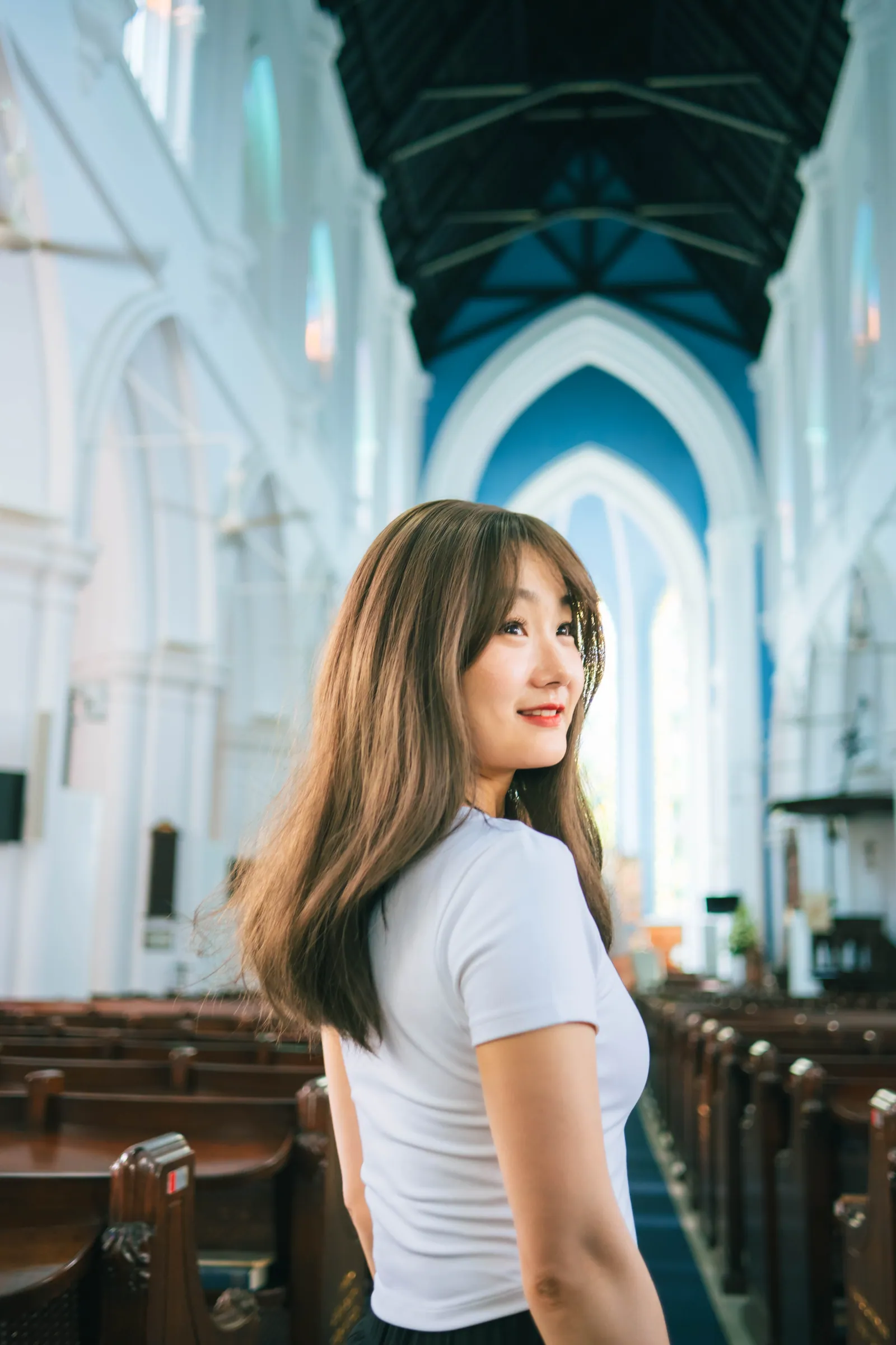 Woman in a white T-shirt looking back over her shoulder in the bright white nave of St Andrew's Cathedral, with wooden pews and a dark timber ceiling